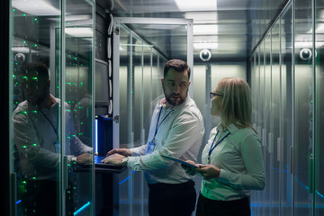 Adult man and woman using tablet and laptop while diagnosing server hardware in modern data center