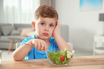 Emotional little boy eating vegetable salad at table in room