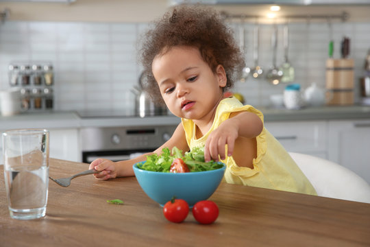 Cute African-American girl eating vegetable salad at table in kitchen