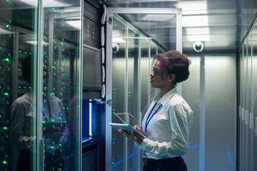 Medium shot of female technician working on a tablet in a data center full of rack servers running...