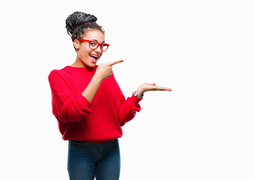 Young braided hair african american girl wearing sweater and glasses over isolated background amazed and smiling to the camera while presenting with hand and pointing with finger.