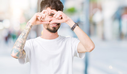 Young handsome man wearing white t-shirt over isolated background Doing heart shape with hand and fingers smiling looking through sign