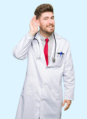 Young handsome doctor man wearing medical coat smiling with hand over ear listening an hearing to rumor or gossip. Deafness concept.