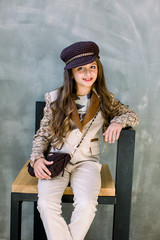 Portrait of adorable smiling little girl in a light suit, a golden sneakers and a hat isolated on a grey sitting on the chair