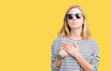 Beautiful young woman wearing sunglasses over isolated background smiling with hands on chest with closed eyes and grateful gesture on face. Health concept.