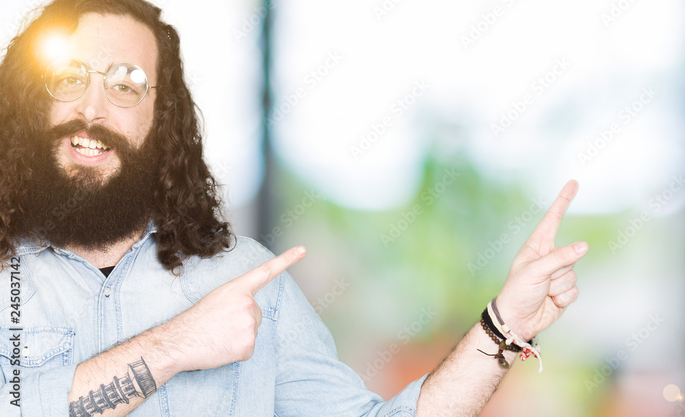 Wall mural Young hipster man with long hair and beard wearing glasses smiling and looking at the camera pointing with two hands and fingers to the side.