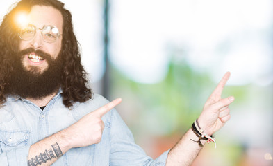 Young hipster man with long hair and beard wearing glasses smiling and looking at the camera pointing with two hands and fingers to the side.