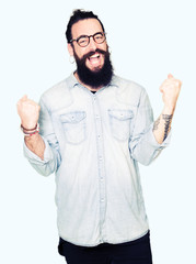 Young hipster man with long hair and beard wearing glasses very happy and excited doing winner gesture with arms raised, smiling and screaming for success. Celebration concept.