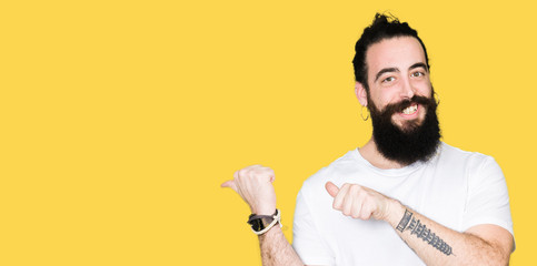 Young hipster man with long hair and beard wearing casual white t-shirt Pointing to the back behind with hand and thumbs up, smiling confident