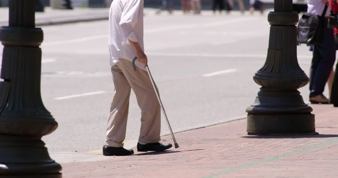 Old man with a cane crosses a city street onto a sidewalk in slow motion