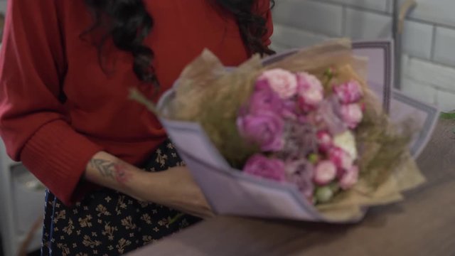 Girl-florist is decorating a bouguet of flowers with decorative paper. She is in flower shop. There also is another flower composition on the table.