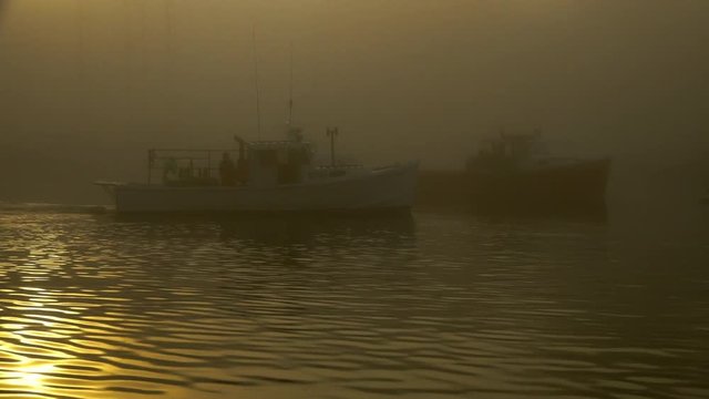 A lobster fishing boat moves quietly through the water and early morning light at sunrise in a foggy and quaint New England harbor.