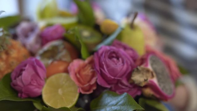 Slow shot of the bouquet in the foreground. Bouguet is very big and non-standart. It include exotic fruits(pineapple,persimmon, kiwi, dragon fruit)and flowers(pink roses).