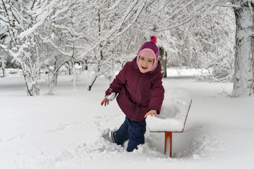 Fototapeta na wymiar Little cute girl in winter outdoors. She shakes off snow from bench. Girl rejoices and fools