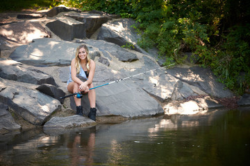 Beautiful blonde young woman fishing near creek wearing coveralls - seated on rock