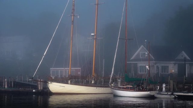 Beautiful vintage sailboats are tied up at a dock in a quaint New England harbor during foggy early morning light.