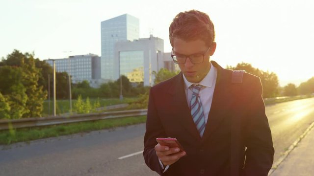 Super slow motion, sun flare, close up young businessman anxiously browsing the internet on his smart phone as he walks down the empty sidewalk on a sunny summer evening