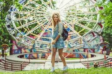young beautiful blond girl in denim overall with a backpack posing in an amusement park, on the...