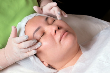 Relaxing and rejuvenating female cosmetological procedure. The hands of a beautician in white gloves apply the cream around the eyes of an Asian girl in a beauty salon or cosmetology center. Close-up.