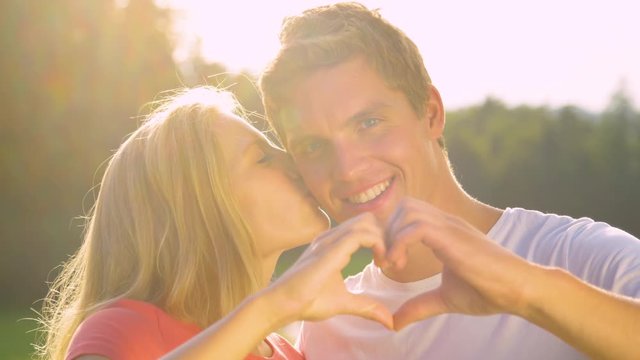 SLOW MOTION, LENS FLARE, CLOSE UP: Beautiful blonde haired woman kisses the smiling Caucasian man on the cheek while holding up a heart shape with their fingers. Young couple enjoying date in nature.