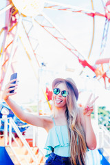 Adorable young girl smiling, showing her tongue and doing selfie with the mobile device at an amusement park. She wears sunglasses and a hat. Portrait of lifestyle