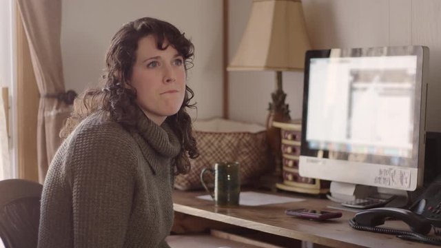 Young woman in sweater works on computer in her home office while listening.