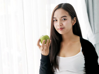 Asian pregnant woman holding green apple near window, lifestyle concept.