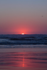 Fototapeta na wymiar Scenic deep red sunset over Pacific Ocean, Oregon Coast. Red sun go down coloring sly and horizon in purple and red colors leaving a thin blurred trace on a wet beach sand. Waves crushing on a beach