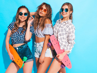Three young stylish smiling beautiful girls with colorful penny skateboards. Women in summer hipster checkered shirt clothes posing near blue wall in studio in sunglasses. Positive models having fun
