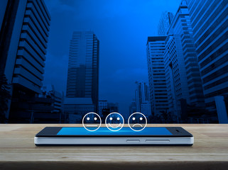 Excellent smiley face rating icon on modern smart mobile phone screen on wooden table over office city tower and skyscraper, Business customer service evaluation and feedback rating concept