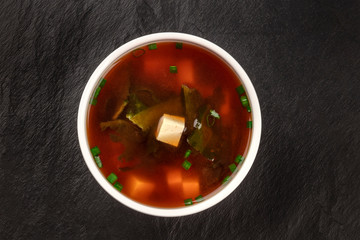 A photo of a bowl of miso shiru soup with tofu, scallions, and wakame seaweed, shot from above on a black background with a place for text