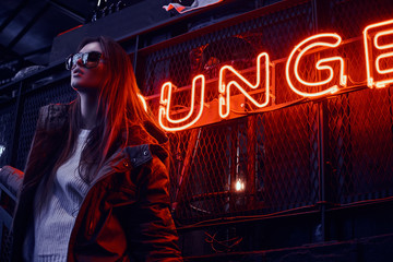 Fototapeta premium Young stylish girl wearing a hoodie coat and sunglasses standing on stairs at underground nightclub with industrial interior