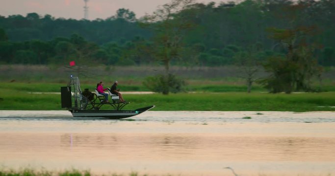 An airboat glides through a central Florida swamp and grassy wetlands in search of wildlife viewing and gator hunting at sunset. Slow motion.