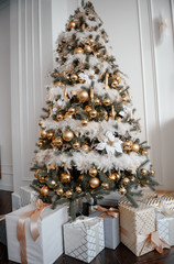 Stylish Christmas living room decor in gold and silver tones. A large beautifully decorated Christmas tree stands in the room next to the modern sofa.