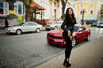 Young curly and sexy woman in leather jacket against red muscle car at street.