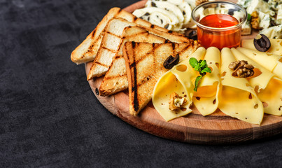 Obraz na płótnie Canvas Cheese plate: Parmesan, cheddar, gouda, mozzarella and other with basil on wooden board on dark background with place for text.Honey and Crackers