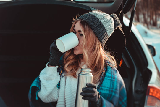 Young Woman Sitting On Car Trunk With Coffee Cups And Thermos In Winter Field