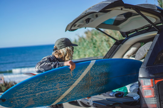 surfer man with long white hair in a cap and sunglasses with a surfboard. returns after surfing, folds the surfboard into the car. Nazare, Portugal.