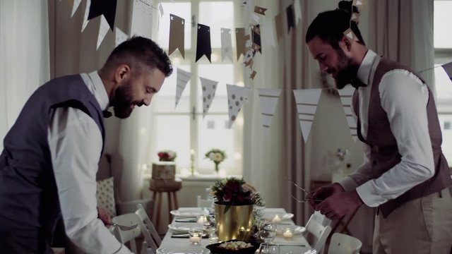 Two men with bows and vests setting a table for an indoor party, talking.