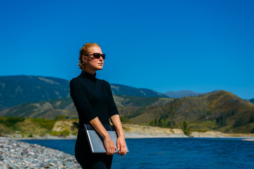 Fototapeta premium Freelancer with laptop on the nature. Blogger hipster traveler business lady pretty girl finished work outdoor. Young blondy woman with sunglasses in black standing next to mountain river