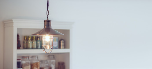 Decorative industrial style metal lamp hanging from the ceiling of a modern kitchen with white walls and shelves with healthy food in the background.