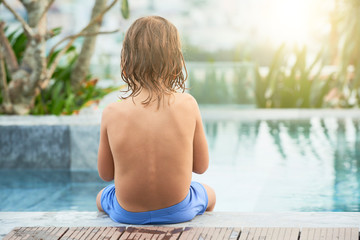 Kid sitting on edge of swimming pool at spa resort, view from the back