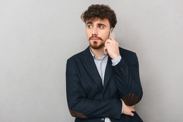 Handsome serious young business man isolated over grey wall background talking by mobile phone.