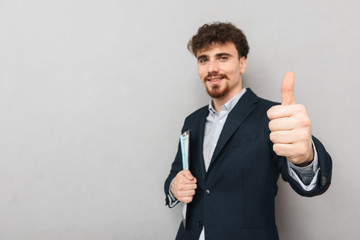 Young business man isolated over grey wall background holding clipboard showing thumbs up.