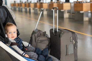 Cute funny caucasian baby boy sitting in stroller near luggage at airport terminal. Child sin...
