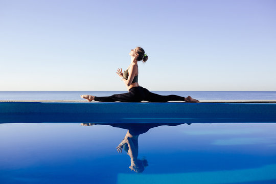 Young attractive woman practicing yoga stretching in One Legged King Pigeon exercise, Eka Pada Raja Kapotasana working out by pool, above beach, relaxing against blue sky. Health and beauty concept