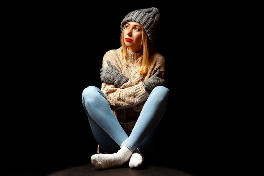 A blonde girl with make-up in a knitted brown sweater and hat with gloves is sitting off her legs crossed on the floor in the studio on a black background and looks up at the imaginary falling snow.
