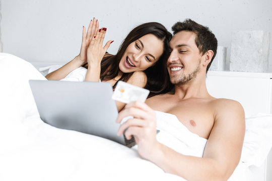 Image of beautiful couple using laptop and credit card, while lying in bed at home or hotel apartment