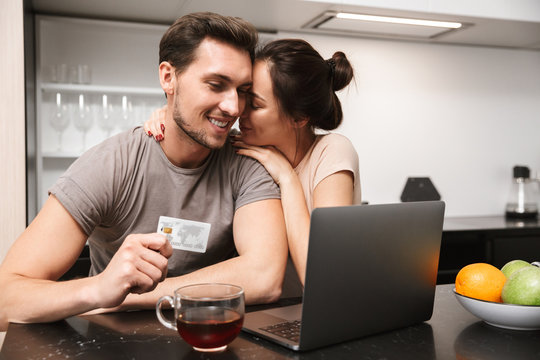 Photo of attractive couple man and woman using laptop with credit card, while sitting in kitchen