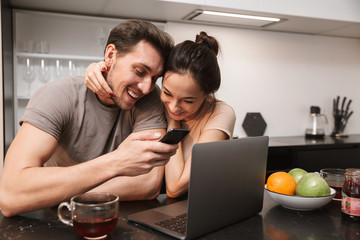Caucasian couple man and woman using laptop with smartphone, while sitting in kitchen